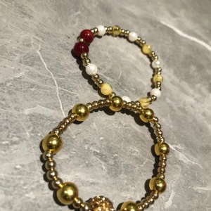 Två pärlarmband i guld och rött - Säljer två eleganta pärlarmband. Ett armband har guld- och silverpärlor med en dekorativ guldfärgad pärla. Det andra armbandet har en mix av vita, röda och guldiga pärlor. Perfekta för att ge en touch av färg och stil. 100kr för båda 50kr för ett
