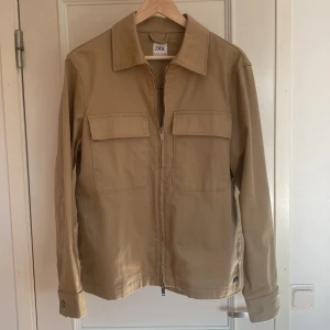 Beige Overshirt från Zara - Säljer en stilren beige overshirt från Zara med dragkedja och två stora fickor framtill. Jackan är perfekt ovanpå en Hoodie eller något liknande men även på en T-shirt. Skick 10/10