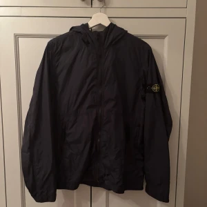 Svart regn/vindjacka från Stone Island - Säljer en stilren mörkblå vindjacka från Stone Island med huva och dragkedja. Jackan har en ikonisk logga på ärmen och är perfekt för blåsiga dagar. Den är lätt och smidig att bära med sig. Passar perfekt för dig som gillar en sportig look. Koster runt 3500 i butik (knappt använd) 160 cm