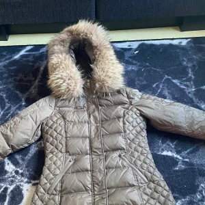 Äkta Rockandblue  - Säljer en snygg brun dunjacka från Rockandblue med quiltad design och pälskantad huva. Jackan har dragkedja och praktiska fickor med dragkedjor. Perfekt för kyliga dagar! Storlek 38 Äkta päls!!  