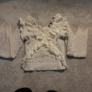Beige stickad tröja från Zara - Säljer en söt beige stickad tröja från Zara med vackra spetsdetaljer och volanger. Tröjan har långa ärmar och en v-ringad design. Perfekt för en romantisk look. 🧶✨