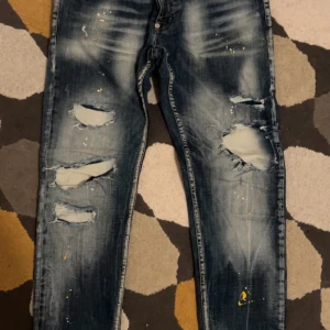 Blå slitna jeans med färgstänk - Snygga blå jeans med slitningar och coola färgstänk. De har en straight passform och är perfekta för en avslappnad stil. Jeansen har en klassisk femficksdesign och knappar i midjan.