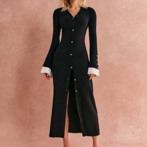 Svart långklänning med knappar - Elegant svart långklänning med knappar hela vägen ner och vita manschetter. Klänningen har långa ärmar och en slits framtill som ger en stilren look. Perfekt för en chic och sofistikerad stil.