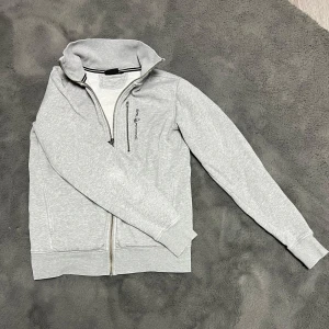 Grå zip-up hoodie från sail racing  - Snygg grå zip-up hoodie från Sail racing med dragkedja framtill och broderad logga på bröstet. Tröjan har långa ärmar och ribbade muddar vid ärmslut och nederkant. Perfekt för en avslappnad stil.