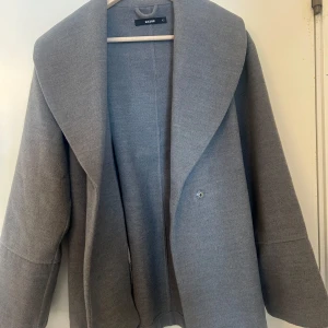 Grå kappa från Bik Bok - Säljer en stilren grå kappa från Bik Bok i storlek XS. Den har en elegant krage och ett bälte i midjan för en snygg passform. Perfekt för kyligare dagar.