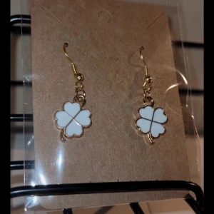 Vita fyrklöver örhängen - Charmiga örhängen med vita fyrklöver i guldtonad ram. Perfekta för att ge en touch av tur och stil till din outfit. De hänger från en enkel krok och är lätta att bära.❤️