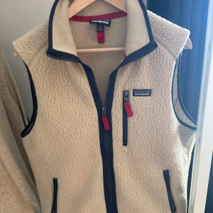 Beige fleeceväst från Patagonia - Säljer en snygg beige fleeceväst frán Patagonia med marinblä detaljer och dragkedjor. Västen har en hög krage och praktiska fickor med röda dragkedjor. Perfekt för lager-pä-lager under kyliga dagar.  Hör av dig vid frågor eller funderingar😁