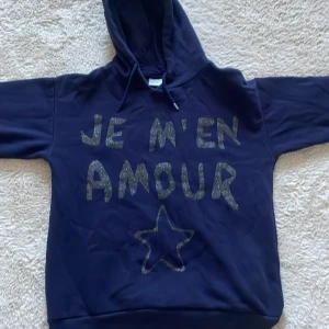 Mörkblå hoodie från Gina Tricot storlek 146-152( SÅLD) - Säljer en mörkblå hoodie från Gina Tricot med texten 'JE M'EN AMOUR' i glittriga bokstäver och en stjärna på framsidan. Perfekt för en avslappnad stil med en mysig huva.