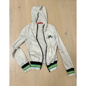 Vit JL vindjacka med huva - Säljer en snygg vit vindjacka med huva från JL. Jackan har en dragkedja framtill och ribbade muddar i svart, grönt och blått vid ärmslut och nederkant. Perfekt för vår och sommar!