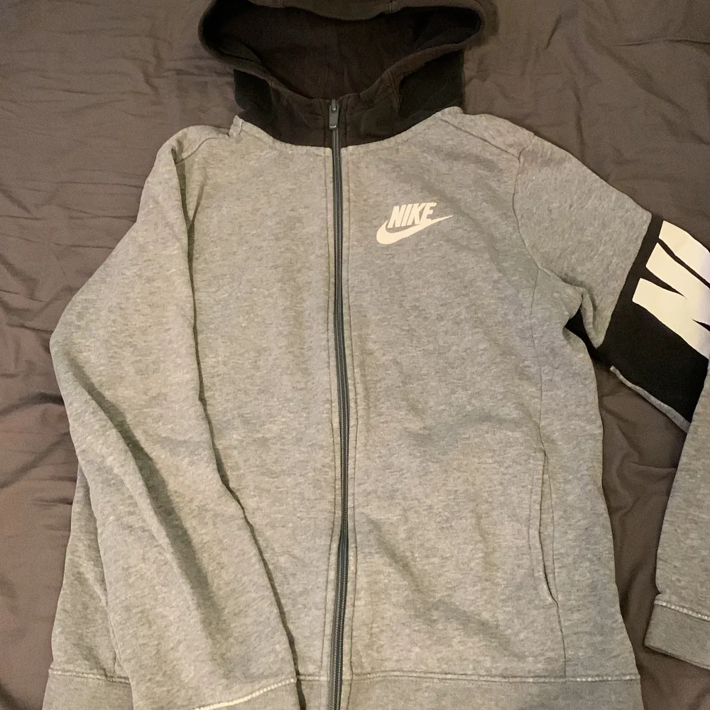 Säljer en grå hoodie från Nike i storlek XS. Den har en svart huva och en dragkedja framtill. Ärmarna är långärmade med ett coolt Nike-tryck på ena ärmen. Perfekt för en avslappnad stil eller träning! Handleden är lite riven. Därav låga priset! . Jackor.