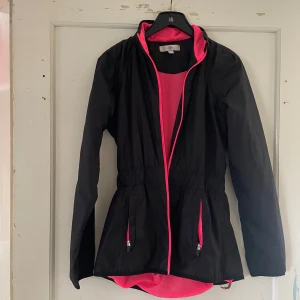 Svart vindjacka med rosa detaljer - Säljer en snygg svart vindjacka med coola rosa detaljer. Jackan har en dragkedja framtill och två praktiska fickor med dragkedjor. Den är perfekt för vår och höst, och har en skön passform med elastisk midja. Insidan är fodrad med rosa mesh för extra ventilation.