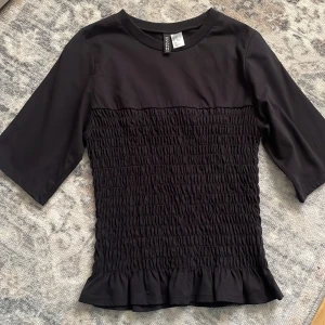 Svart topp från H&M Divided - Snygg svart topp från H&M Divided med trekvartsärm och smockdetaljer på kroppen. Toppen har en rund halsringning och en liten volangkant nedtill. Perfekt för en stilren look.