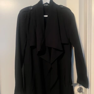 Svart trenchcoat - Elegant svart trenchcoat med långa ärmar och knappar på axlarna. Perfekt för en stilren look under höst och vår. Den har en klassisk design med en draperad krage.