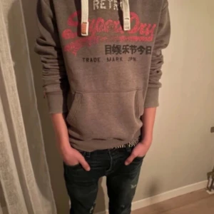 Grå hoodie från Superdry - Säljer en grå hoodie från Superdry med röd logga och text på framsidan. med vita snören och en stor magficka. I storlek XS  säljer då för 249 priset kan diskuteras 