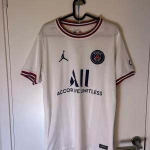 Vit PSG fotbollströja med Jordan-logga - Snygg vit PSG fotbollströja med korta ärmar och Jordan-logga. Tröjan har röda och blå detaljer på krage och ärmslut samt PSG-emblem på bröstet. Perfekt för fotbollsfans!