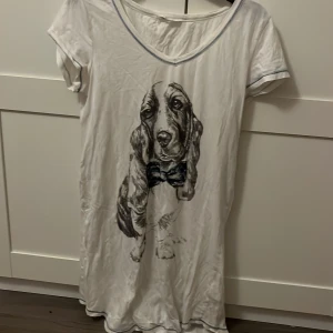 Vit t-shirtklänning med hundmotiv från Lindex - Säljer en vit t-shirtklänning från Lindex med ett stort tryck av en hund med fluga på framsidan. Klänningen har korta ärmar och en v ringad med blå söm. Man kan ha det som nattlinne här själv inte använt