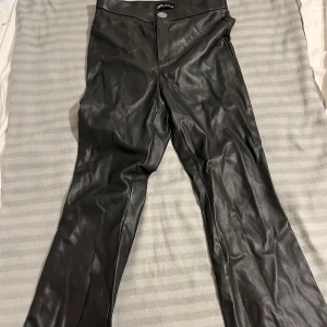 Svarta skinnbyxor från Zara - Snygga svarta skinnbyxor från Zara med hög midja och dragkedja. Perfekta för en stilren look med en modern touch. Byxorna har en knappdetalj vid midjan och är tillverkade i ett syntetiskt material som ger en läderliknande finish. De har använts få tal gånger!