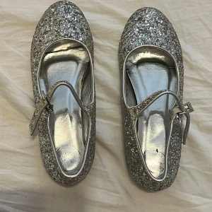 Glittriga silverfärgade ballerinaskor - Snygga ballerinaskor i silver med glittrig yta och justerbart spänne. Perfekta för att lysa upp vilken outfit som helst. Använda några gånger