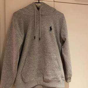 Grå hoodie från Ralph Lauren - Säljer en stilren grå hoodie från Ralph Lauren med klassisk logga på bröstet. Tröjan har en justerbar huva med snören och en praktisk magficka. Perfekt för en avslappnad stil.