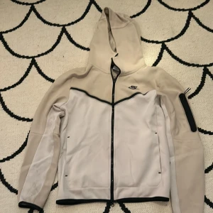 Beige och vit tech fleece från Nike - Snygg beige och vit hoodie från Nike med dragkedja och Luvva . Hoodien har långa ärmar och en praktisk ficka på ärmen. Perfekt för en vanlig dag.Den har lite fläckar vid fickorna men syns bara när man kollar riktigt noga.