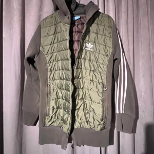 Grön quiltad jacka från Adidas - Snygg grön quiltad jacka från Adidas med huva och dragkedja. Jackan har långa ärmar med de klassiska tre ränderna i vitt längs armarna. Grönare än bilderna. Stl ca M, står xxl på lappen.