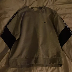 Grå sweatshirt från Lacoste - Snygg grå sweatshirt från Lacoste med svarta detaljer på ärmarna och logotyp längs sidorna. Perfekt för en avslappnad stil. Klassisk rund hals och ribbade muddar.