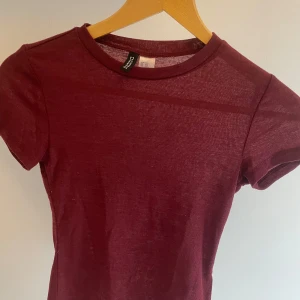 Vinröd t-shirt från hm - Snygg vinröd t-shirt från Divided i en mjuk bomullsmix. Den har en klassisk rund halsringning och korta ärmar, perfekt för en avslappnad stil. Den är lite genomskinlig och har lite intimissmi vibe. Är i st xxs men passar mig som har s/xs
