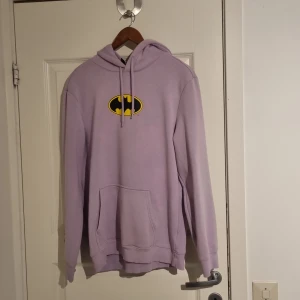 Lila Batman-hoodie - Cool lila hoodie med Batman-logga på framsidan och serietidningstryck på baksidan. Den har en klassisk känguruficka och justerbar huva.