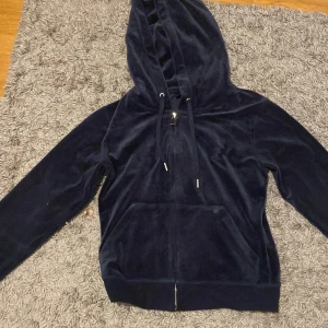 Mörkblå velour hoodie - Snygg och bekväm mörkblå hoodie i mjukt velourmaterial. Den har en dragkedja framtill och en broderad detalj på bröstet. Perfekt för en avslappnad stil.