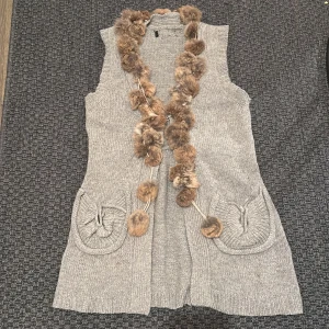 Grå stickad väst med fuskpälsdetaljer - Snygg grå stickad väst med mjuka fuskpälsdetaljer längs kanten. Västen har två praktiska fickor framtill och en elegant, lång design. Perfekt för att lägga till lite extra stil till din outfit.