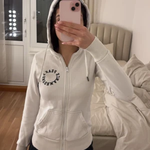 Vit hoodie med dragkedja från Peak Performance - Säljer en vit hoodie från Peak Performance med dragkedja och justerbar huva. Den har ribbade muddar och en logotyp på bröstet samt huvan. Perfekt för en avslappnad stil.