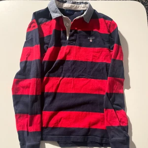 Randig pikétröja från GANT - Snygg randig pikétröja från GANT i rött och blått med klassisk krage och knappar. Perfekt för en avslappnad stil med en touch av elegans.