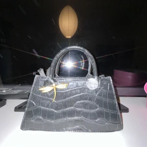 Svart handväska med krokodilmönster - Snygg svart handväska med krokodilmönster och en dekorativ guldfärgad trollslända. Väskan har två handtag och en knappdetalj med zebramönster. Perfekt för att ge en elegant touch till din outfit.