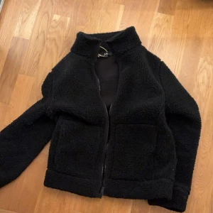 Svart teddyjacka från C&A - Mysig svart teddyjacka från C&A i storlek XS. Jackan har en dragkedja framtill och är långärmad. Perfekt för kyliga dagar när du vill hålla dig varm och bekväm.