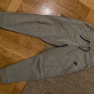 Grå mjukisbyxor från Nike - Säljer ett par grå mjukisbyxor från Nike med snörning i midjan och elastiska muddar vid bensluten. Perfekta för en avslappnad stil eller träning. Byxorna har en liten Nike-logga på sidan.