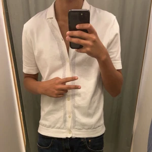 Vit kortärmad skjorta Från EAST WEST - Snygg vit kortärmad skjorta med knappar och subtila randiga detaljer längs knäppningen. Perfekt för en stilren och avslappnad look. Org pris 799kr