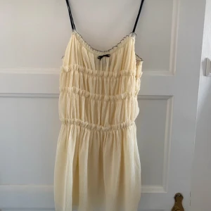 Beige klänning från Zara - Säljer en söt beige klänning från Zara med smala axelband och volangdetaljer. Klänningen har en liten svart rosett framtill och är perfekt för varma dagar.