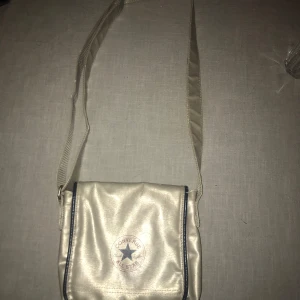 Beige/silver axelväska från Converse - Snygg beige/silver axelväska från Converse med justerbar axelrem. Väskan har ett stort fack med dragkedja och en praktisk framsida med logotyp. Perfekt för att bära med sig det nödvändigaste.