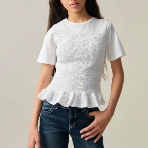 Vit topp med volang från Gina Tricot - Säljer en söt vit topp från Gina Tricot med korta ärmar och volangdetalj vid nederkanten. Toppen har en smockad design framtill som ger en fin struktur. Perfekt för en stilren look!