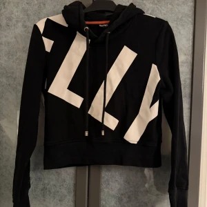 Svart och vit hoodie från Elly Pistol - Snygg svart croppad hoodie från Elly Pistol med stora vita bokstäver som dekoration med deras logga. Tröjan har en justerbar huva med dragsko och långa ärmar. Perfekt för en sportig look eller avslappnad stil. Stl XL men är mycket liten i storlek. Motsvarar mer en M, därav har jag även skrivit det i annonsen.