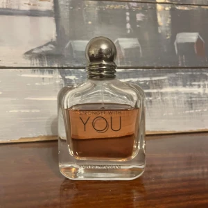 Stronger With You parfym från Emporio Armani 100ml - Elegant parfymflaska med en rundad silverfärgad kork. Doften är varm och kryddig, perfekt för den som vill ha en sofistikerad och modern touch. Flaskan är genomskinlig med en ljus bärnstensfärgad vätska inuti.