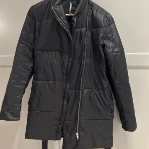 Svart pufferjacka från Whyred - Snygg svart pufferjacka från Whyred med dragkedja och bälte i midjan. Jackan har en quiltad design och långa ärmar, perfekt för kyligare dagar. Stilren och enkel att matcha med olika outfits.