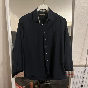 Burberry Skjorta - Snygg mörkblå skjorta från Burberry med klassiska rutiga detaljer på ärmsluten och i kragen. Skjortan har långa ärmar och knappar framtill. Perfekt för en stilren look. Skriv för intresse🤩
