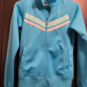 Blå träningsjacka från Nike - Snygg blå träningsjacka från Nike med dragkedja och långa ärmar. Jackan har ljusgrön och vita ränder på bröstet och en liten Nike-logga. Perfekt för träning eller en sportig look.