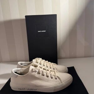 Vita Saint Laurent skor med text på sula. - Vita Saint Laurent-sneakers med text på sulan. Skorna är i fint skick, bedömt som 8/10, med bara lätt användning. Perfekt val för dig som vill ha en snygg och stilren sko med lyxig känsla. Pris: 2499 kr.