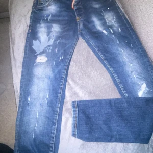 Blå DSQ Ibrahimovic icon - Säljer ett par coola blå jeans med slitningar och en tryckt detalj på benet. De har en klassisk femficksdesign och en bekväm passform. Perfekta för en avslappnad stil. Blå dsqaured Ibrahimovic icon