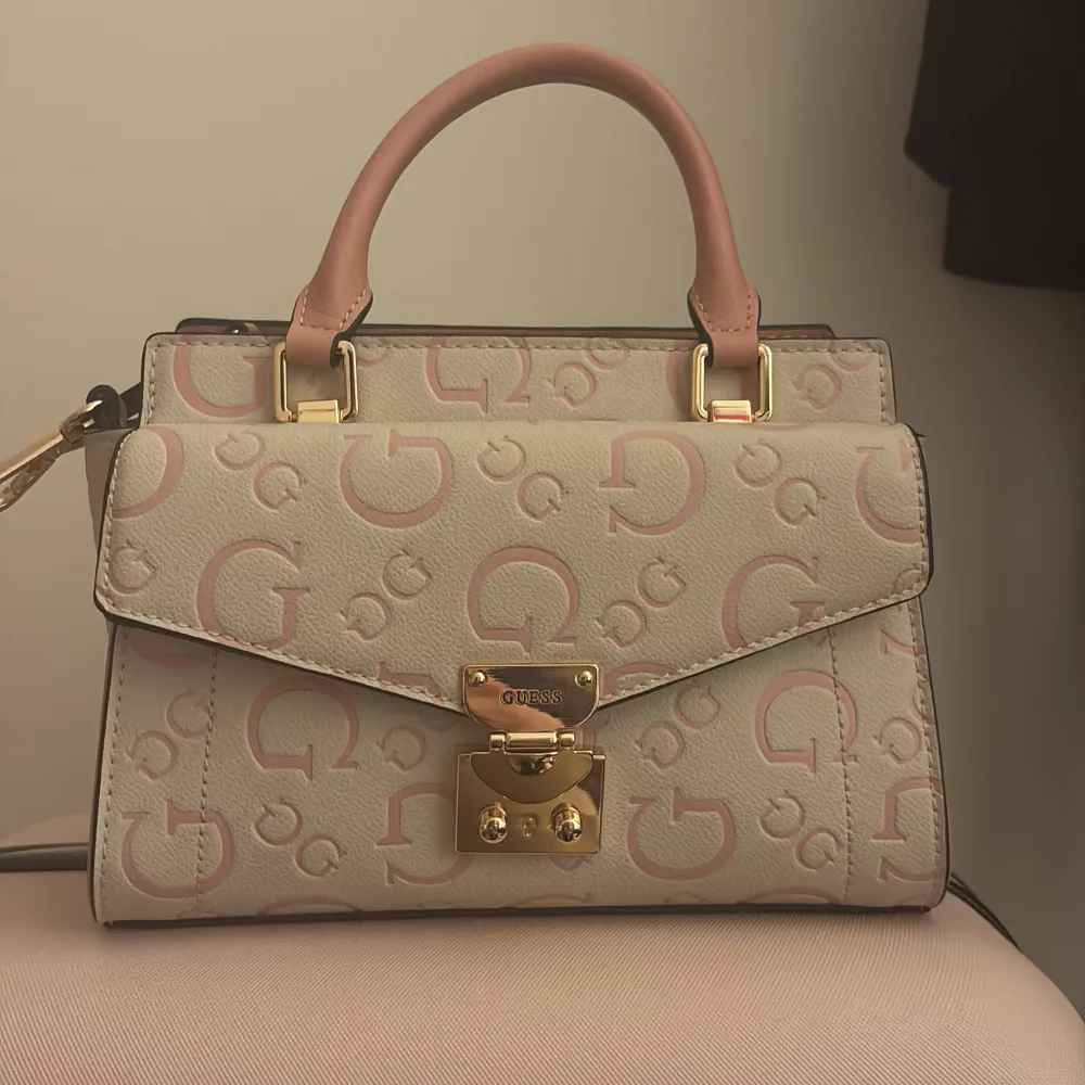 Säljer en snygg handväska från Guess i beige med rosa detaljer och ett stort G-mönster. Väskan har ett elegant handtag i rosa och en guldfärgad låsdetalj framtill. Väskan är använd 1 gång, den är i nytt skick . Väskor.