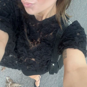 Svart klänning med blommiga detaljer - Säljer en elegant svart blus med blommiga detaljer i ett genomskinligt material. Perfekt för en utekväll eller fest. Blusen  har korta ärmar och en tight passform som framhäver figuren. Den är både stilren och trendig, vilket gör den till ett måste i garderoben! Rensar garderoben, därav priserna!