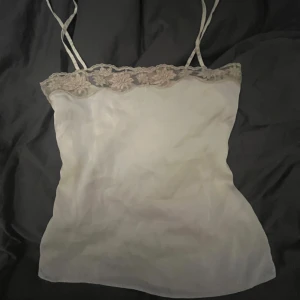 Vit linne med spetsdetaljer - Supergulligt vitt linne med tunna axelband och vacker blommönstrad spetsdetalj längs kanten (ser beige ut på bild men är ljusrosa). Linnet är i bra skick och använts en gång!! Finns ingen lapp så vet ej var den är ifrån eller vilken storlek men skulle säga att den passar S-XS 