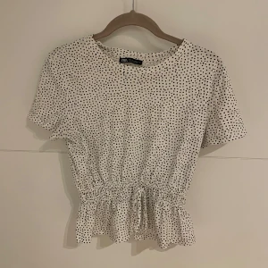 Vit prickig topp från Zara - Säljer en söt vit topp med svarta prickar från Zara. Den har korta ärmar och en smickrande knytning i midjan som ger en fin volangdetalj. Perfekt för både vardag och fest!